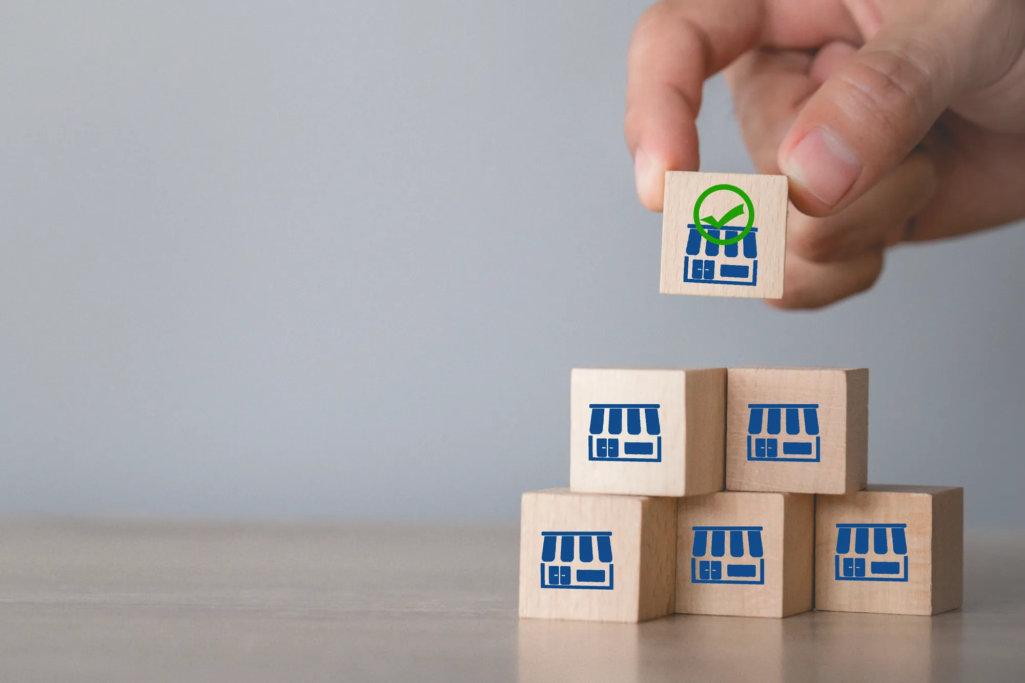 Hand placing a wooden block with a storefront icon and green checkmark atop a stack of matching storefront blocks, symbolizing consistent, on-brand locations in franchise marketing