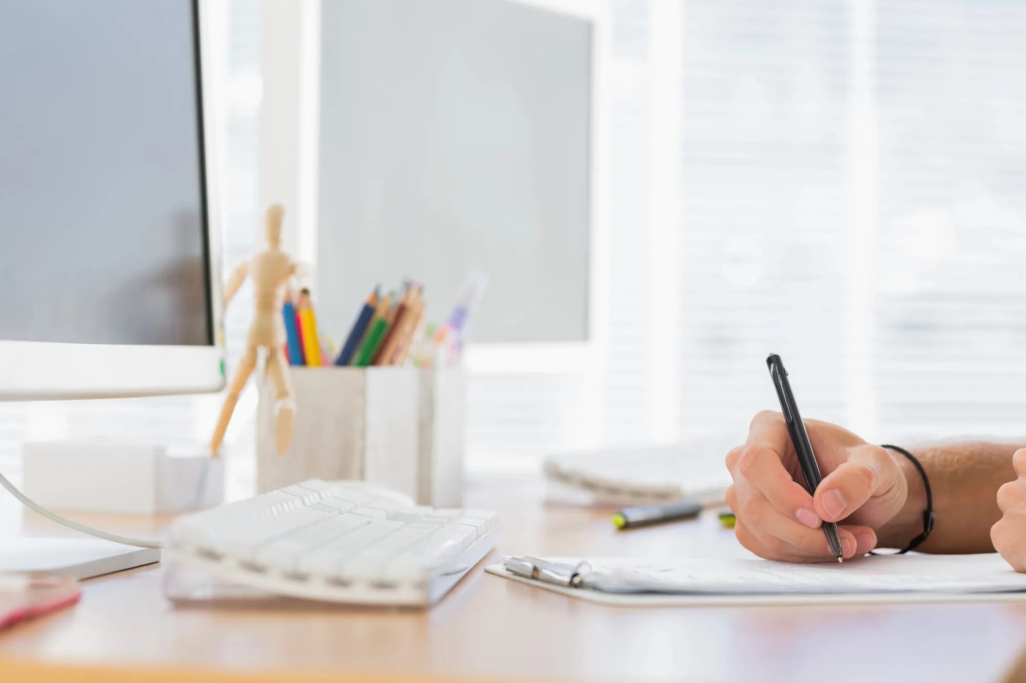 Hand writing notes on a clipboard at a bright desk with a computer monitor and colored pencils, representing budget planning when weighing SEO vs PPC