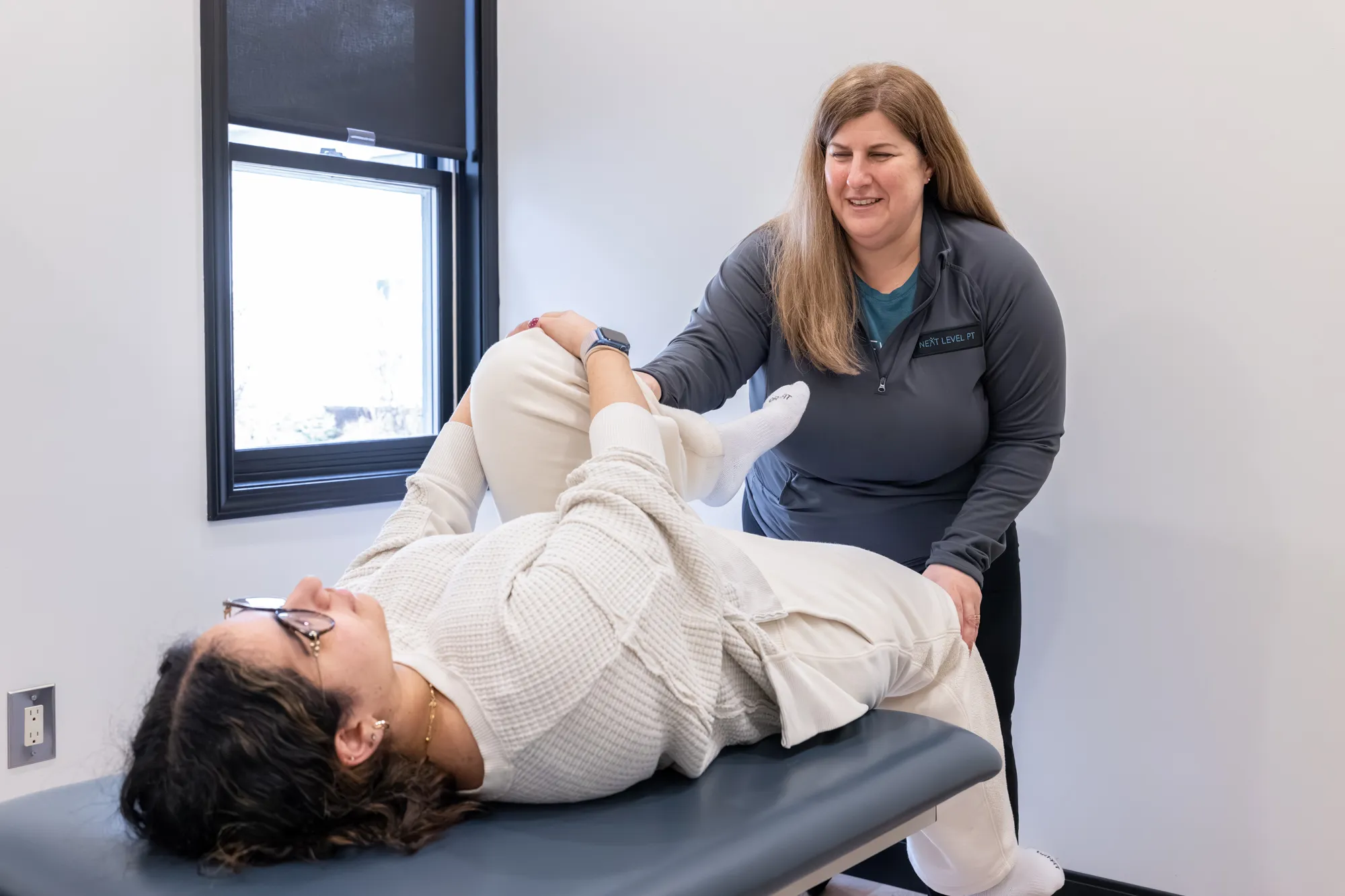 Physical therapist performing knee stretch on a woman lying on an exam table in a clinical room.