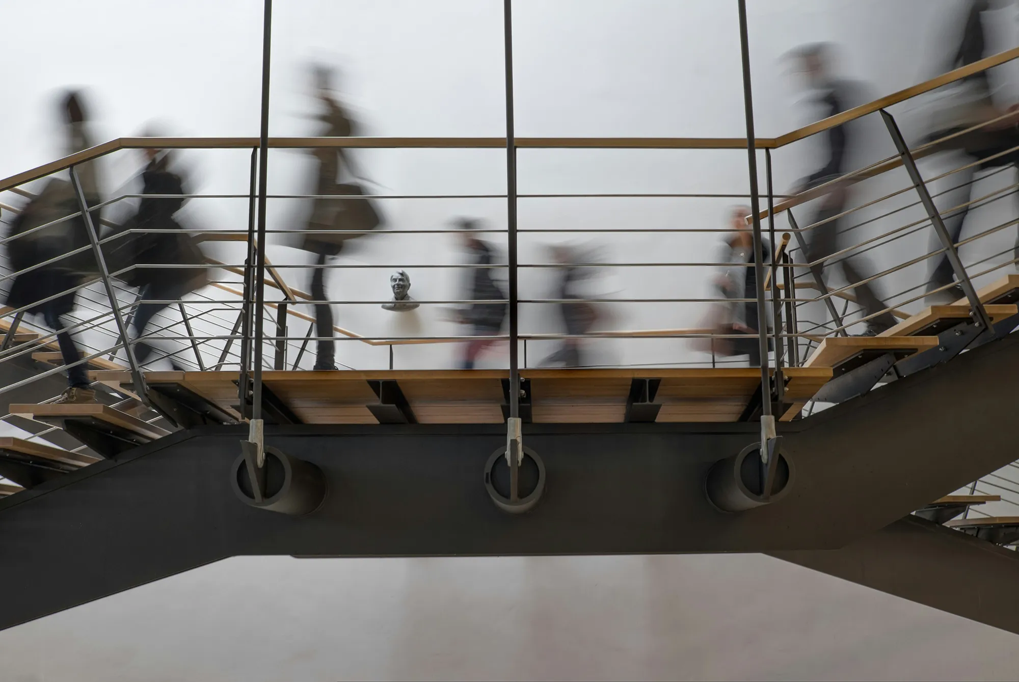 Blurred people walking up and down a modern staircase with a bust sculpture on the wall behind.