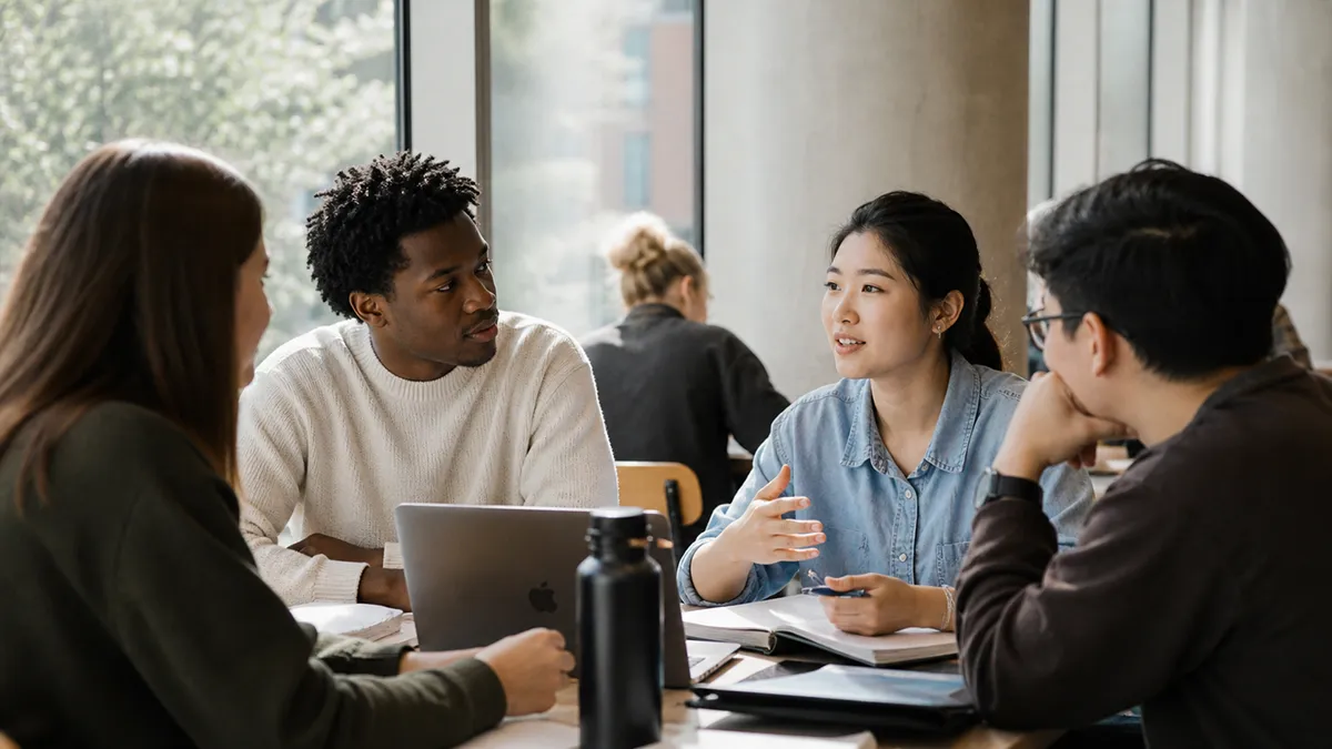 Four diverse students engaged in discussion with open books and laptop in a bright study room.