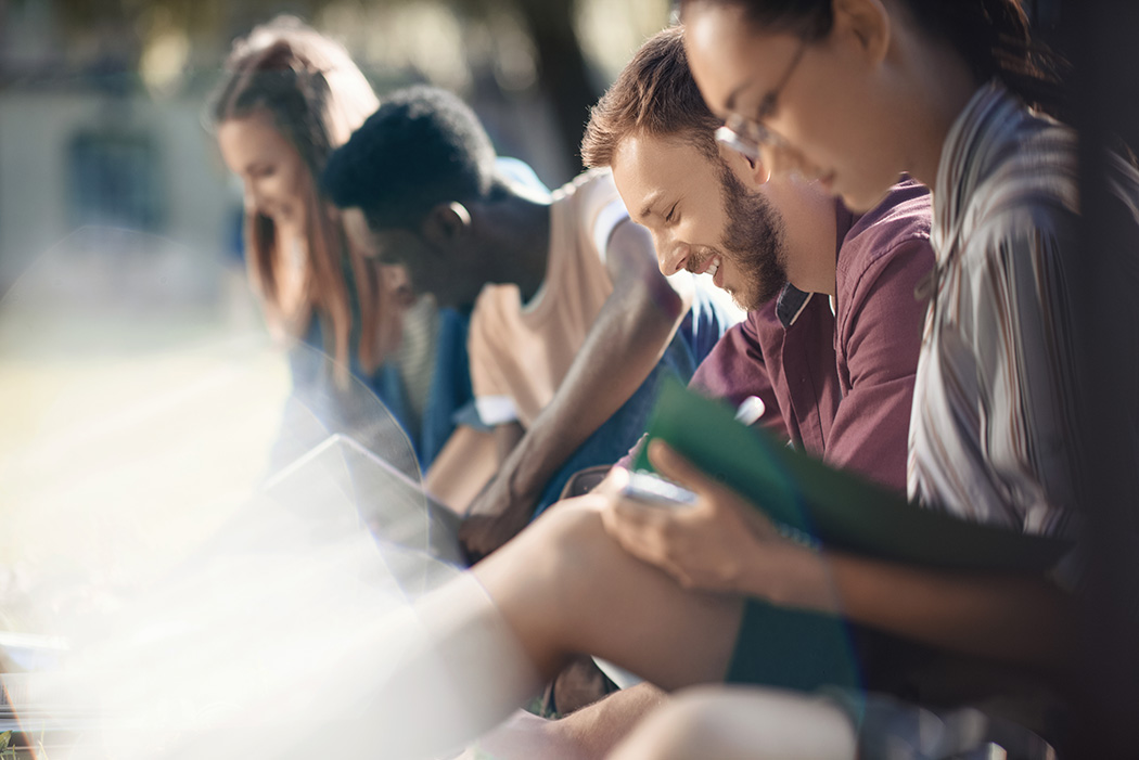 Four young adults sitting outdoors reading books and smiling on a sunny day.