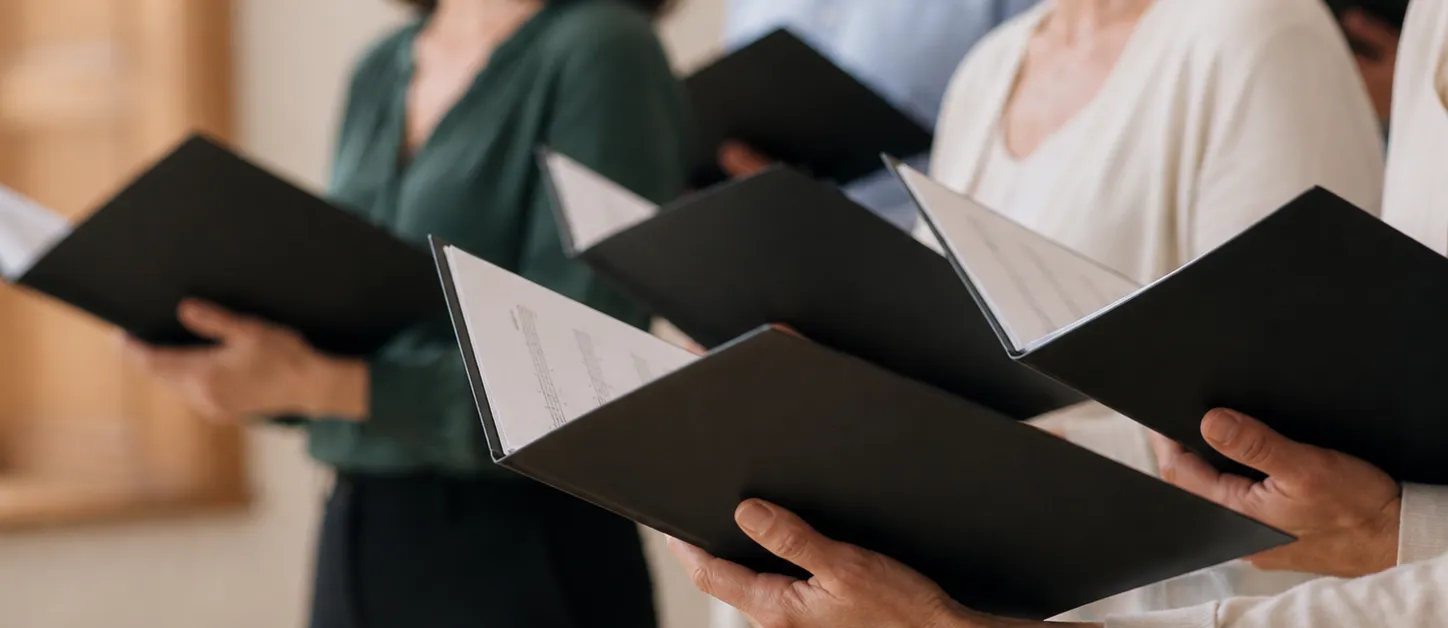 Group of people holding black folders with sheet music, appearing to sing in a choir practice.