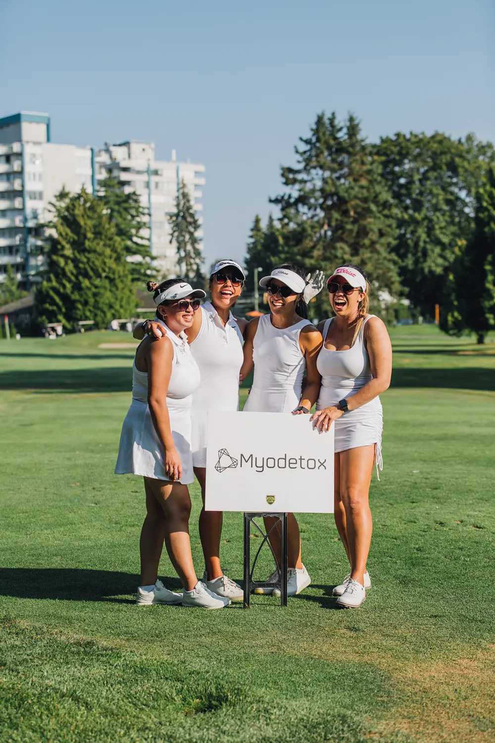 Four women in white golf outfits smiling and posing around a sign that says Myodetox on a golf course.