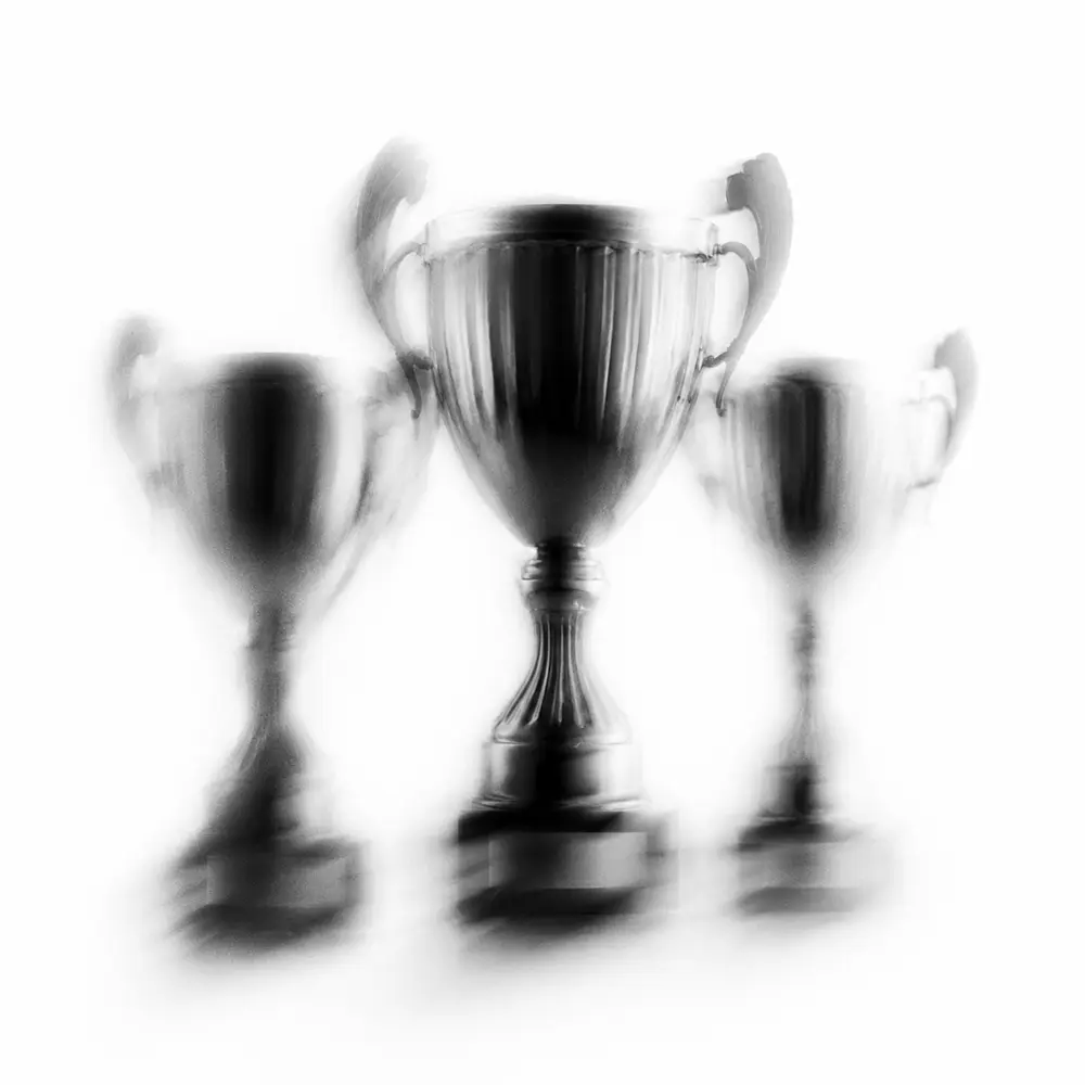 Three blurred silver trophies with handles on a white background, the center one larger than the others.