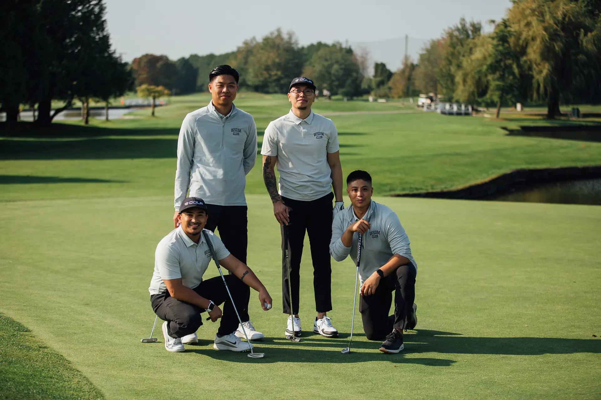 Four men on a golf course green wearing WSDM CLUB shirts, two crouching with golf clubs and two standing.