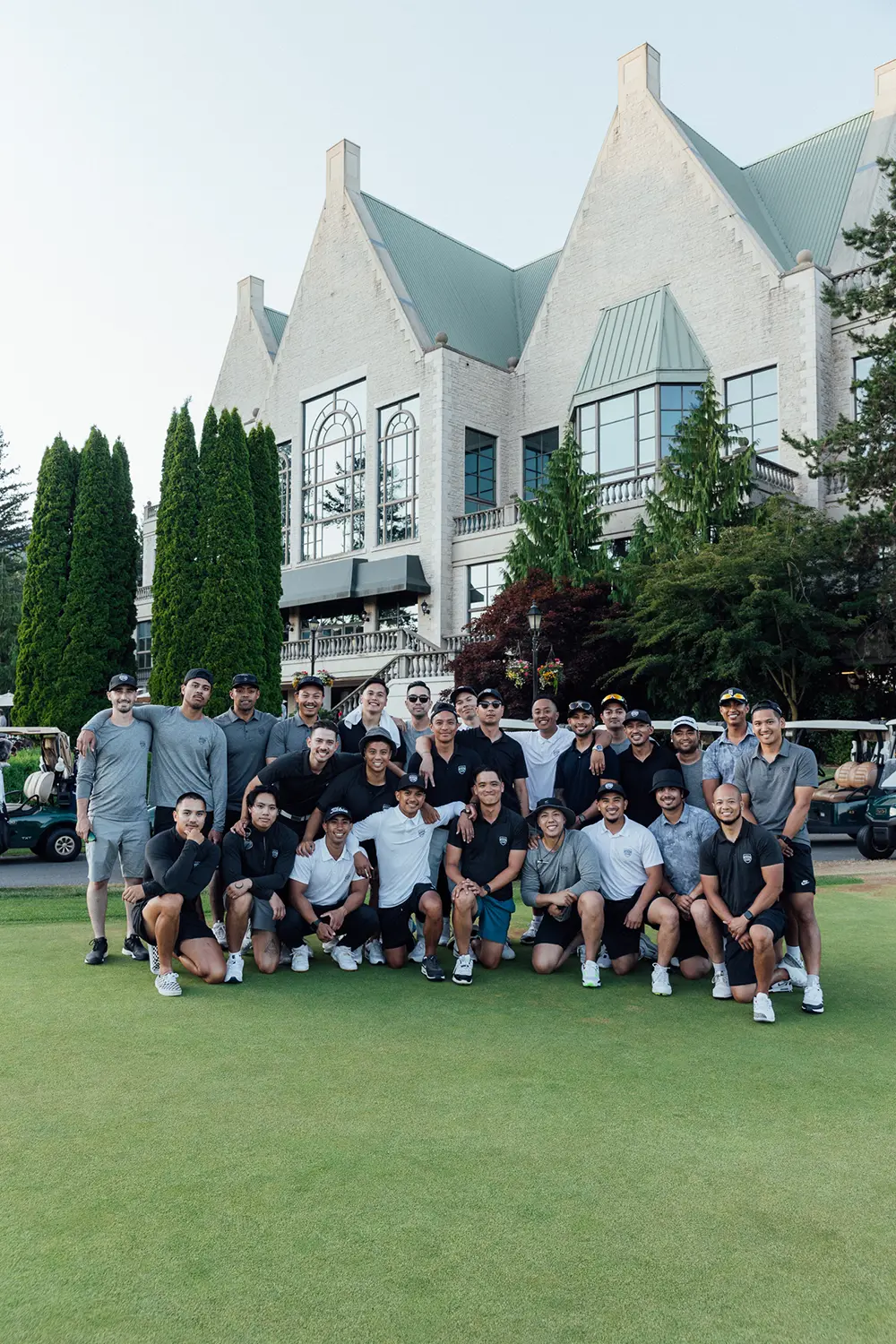 Group of smiling men posing on green lawn in front of a large building with tall windows and greenery.