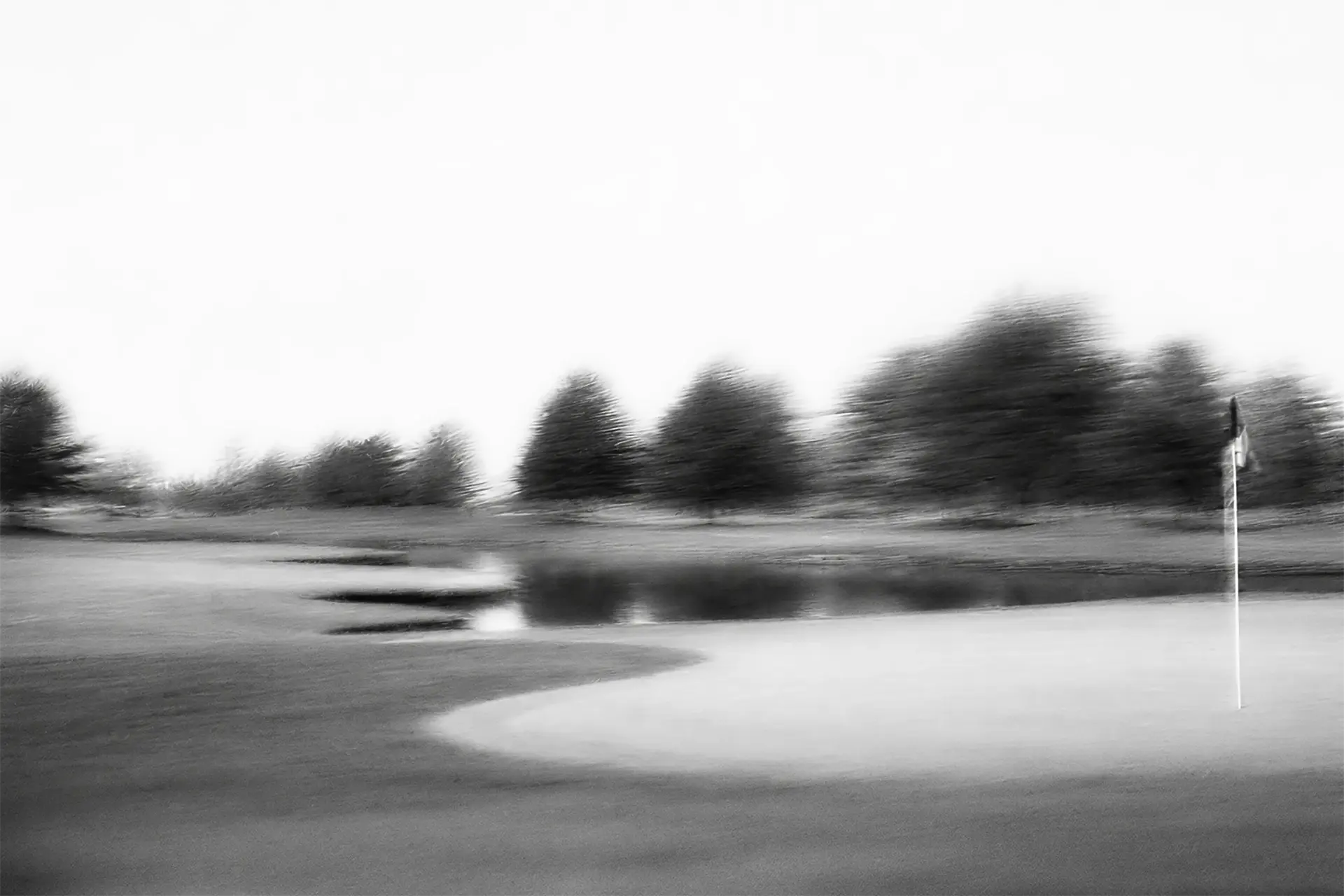 Blurry black and white photo of a golf green with a flagstick and trees in the background.