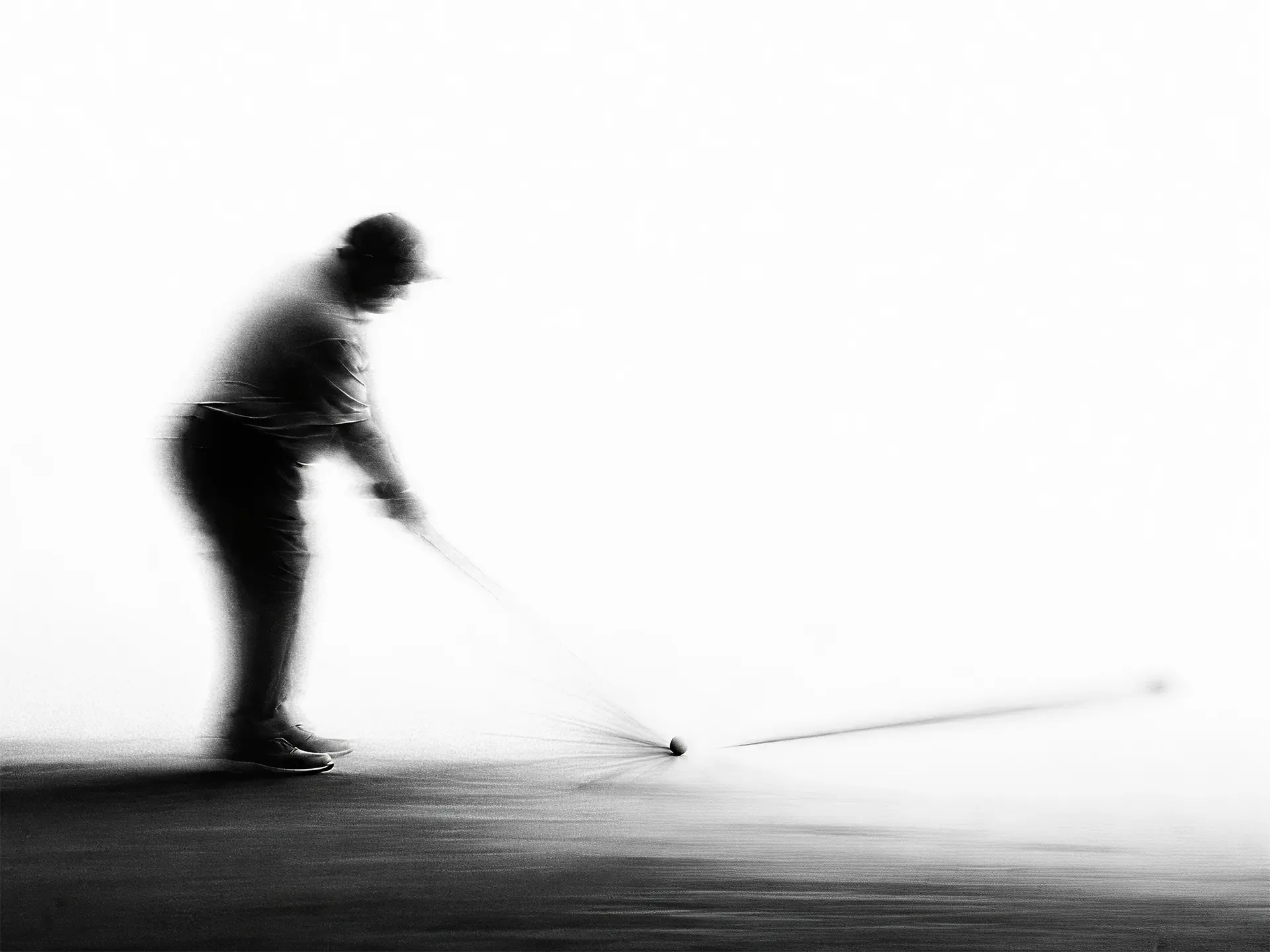Blurred black and white image of a golfer swinging toward a ball on the ground.