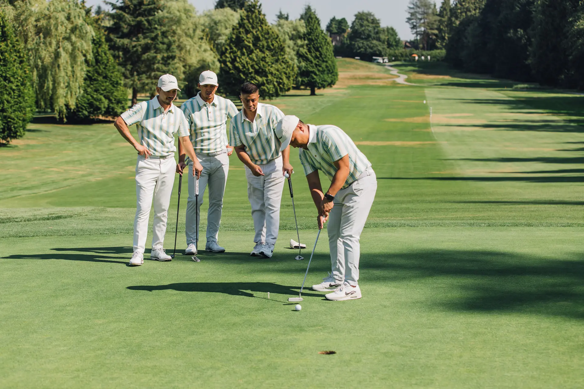 Four men in matching shirts golfing on a green course, one putting while others watch.