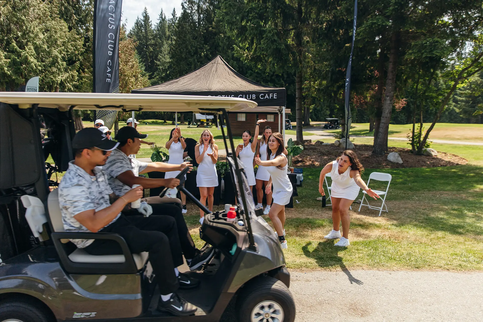 Two men driving a golf cart past women in white dresses cheering at a golf course event.