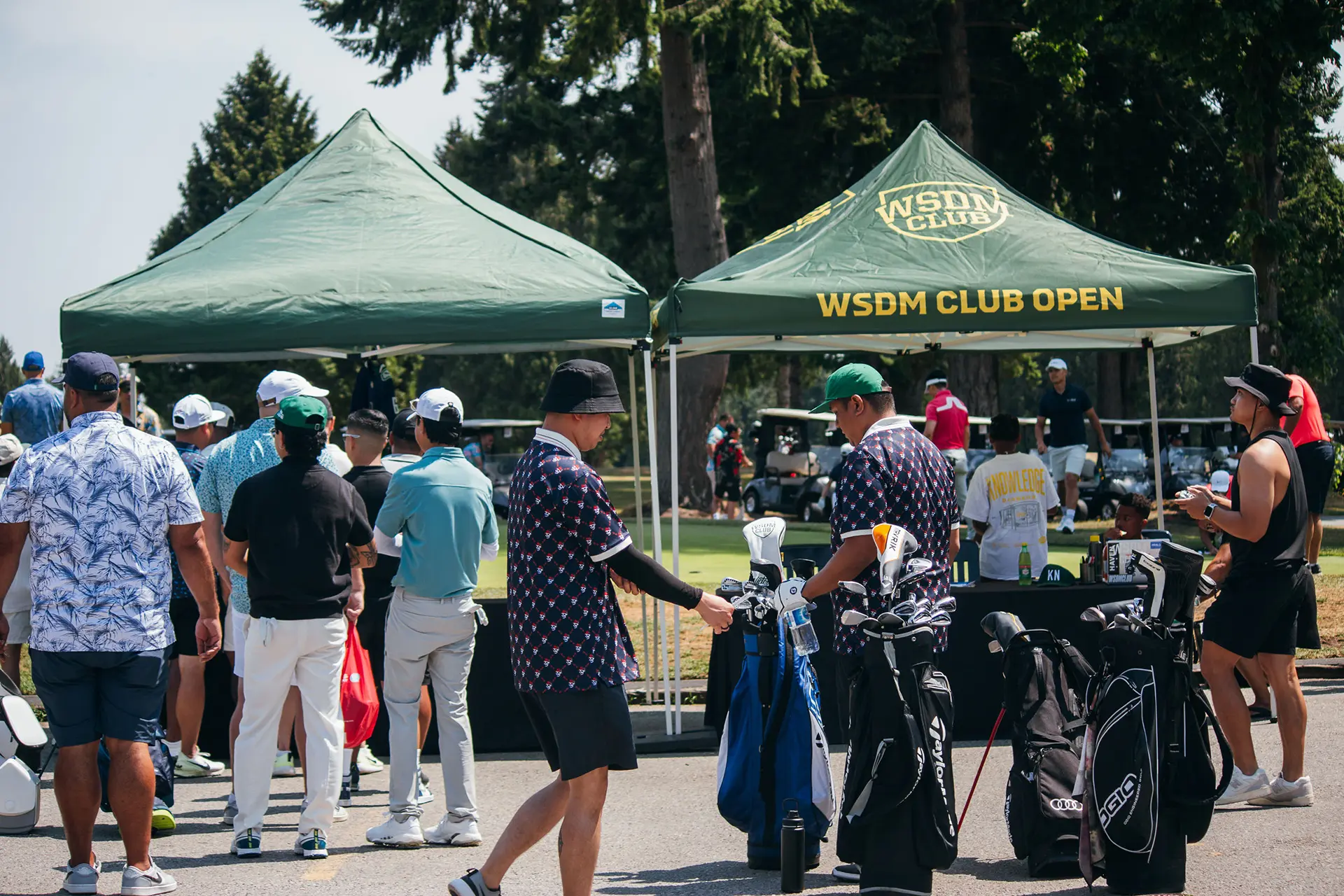 People gather at two green tents with golf bags and clubs at an outdoor WSDM Club Open event.
