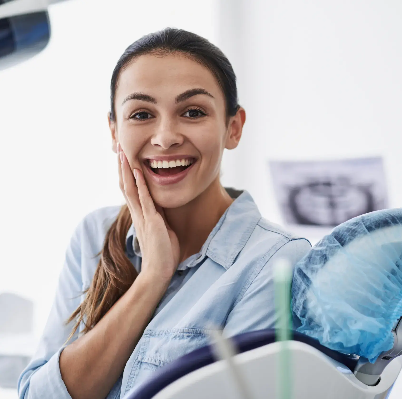 Smiling young woman sitting in a dental chair, touching her cheek, with dental equipment nearby.