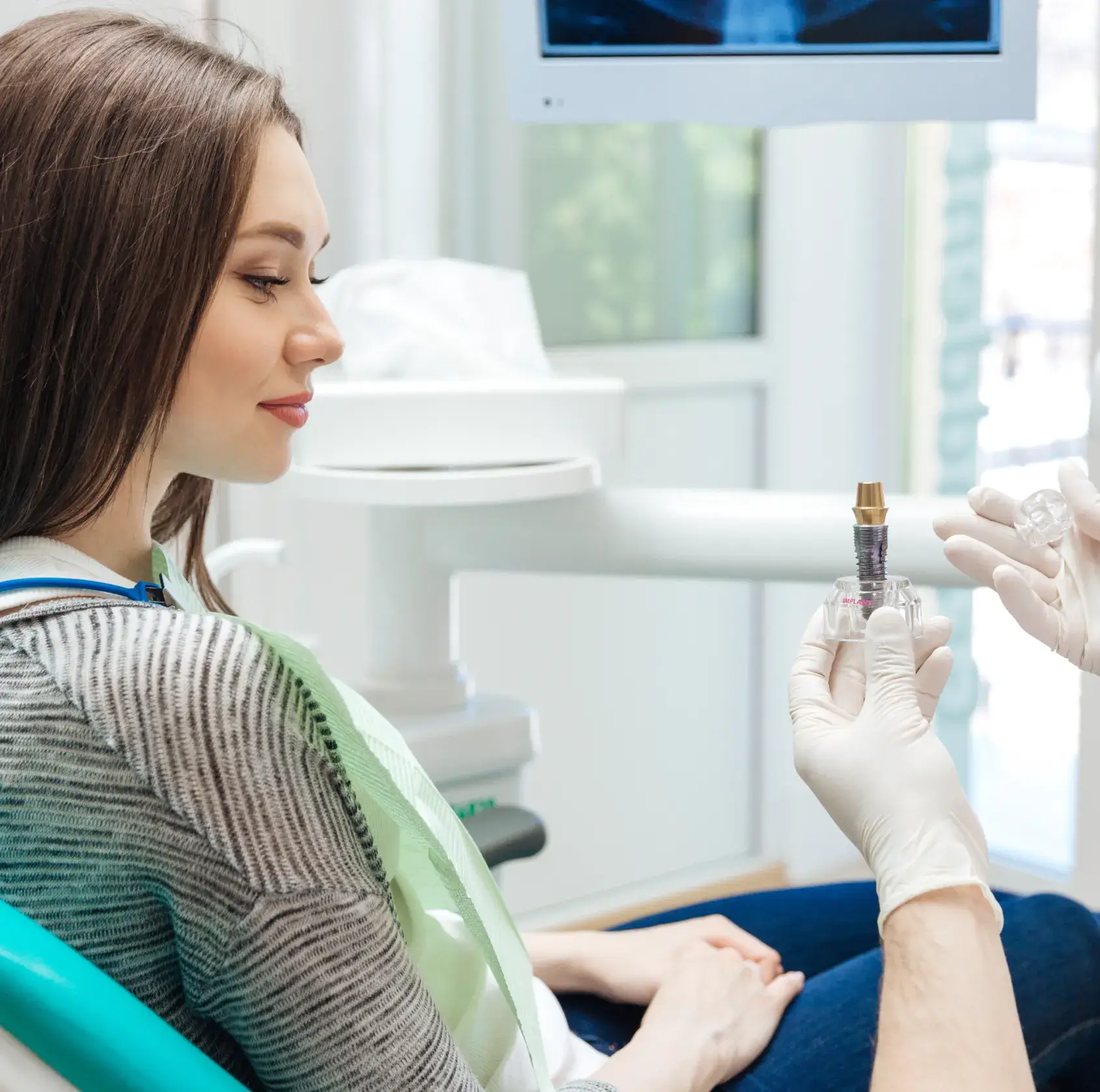 Dentist showing dental implant model to a seated female patient in a dental clinic.