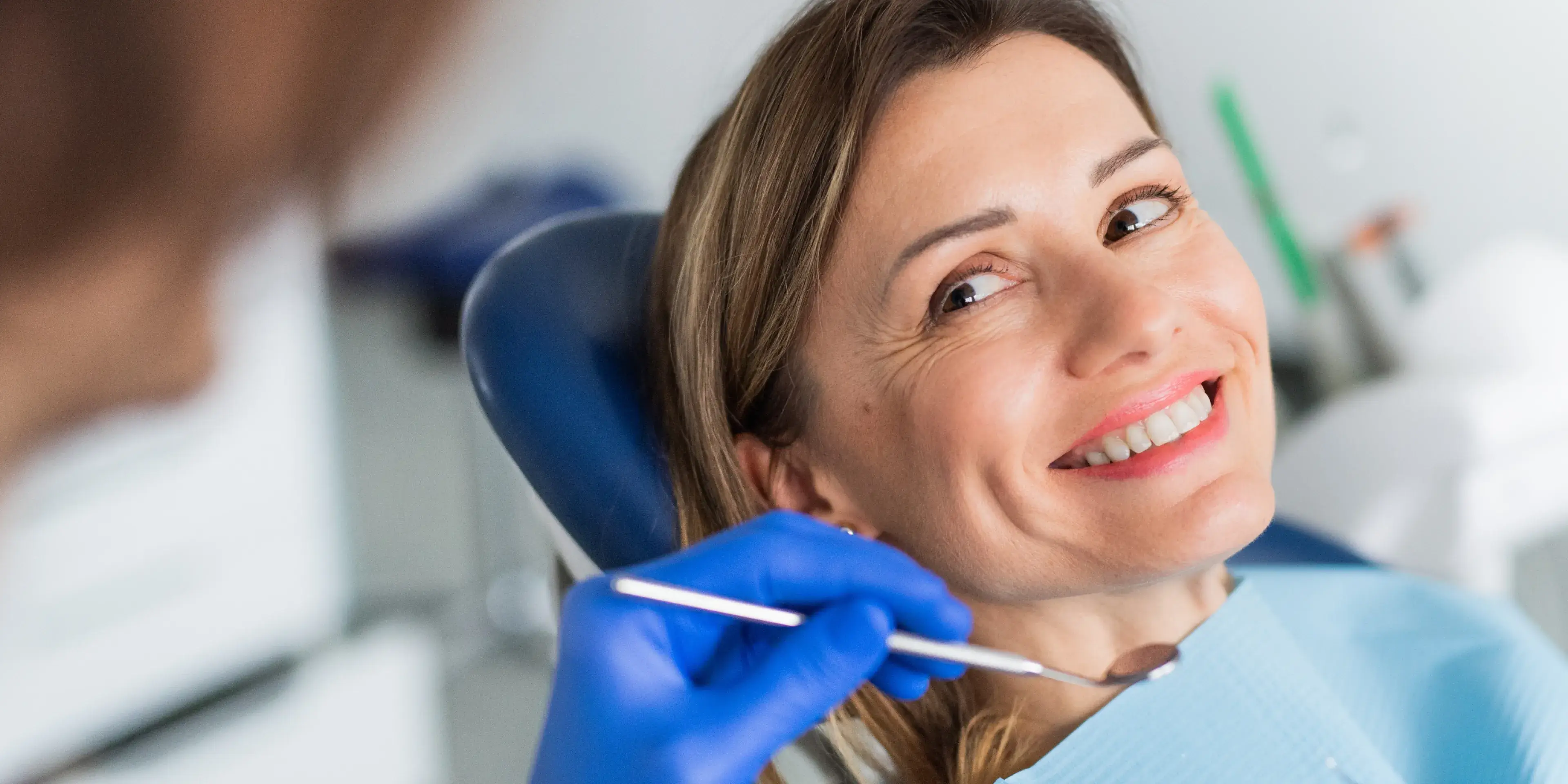 Smiling woman in dental chair receiving checkup from dentist with blue gloves holding a dental mirror.