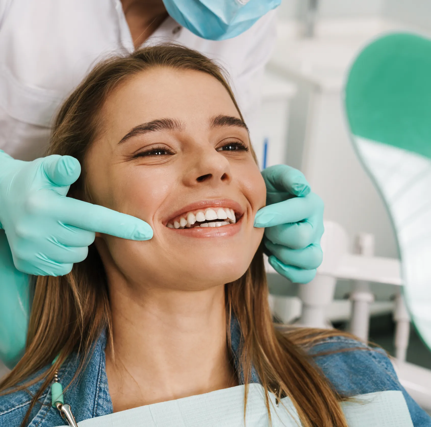 Dentist wearing gloves pointing at a smiling young woman's teeth during a dental exam.
