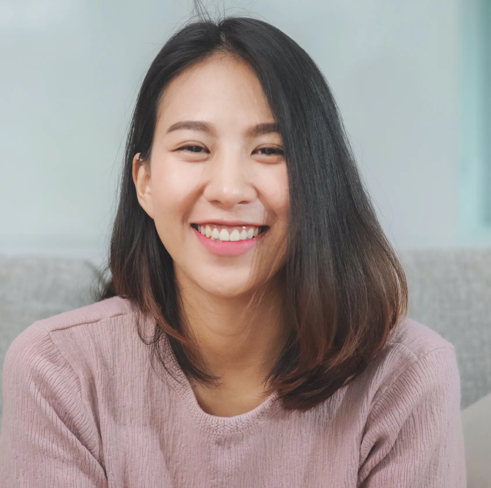 Young woman with dark hair smiling brightly, wearing a light purple sweater indoors.