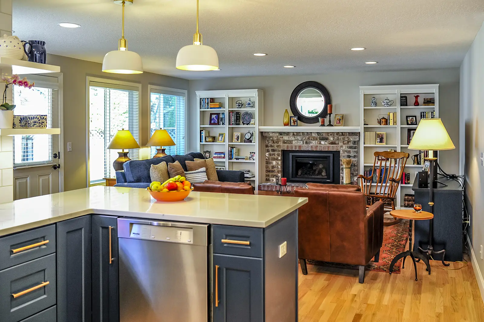 Open living room with a blue sofa, brown chairs, white shelves, fireplace, and kitchen island with fruit bowl.