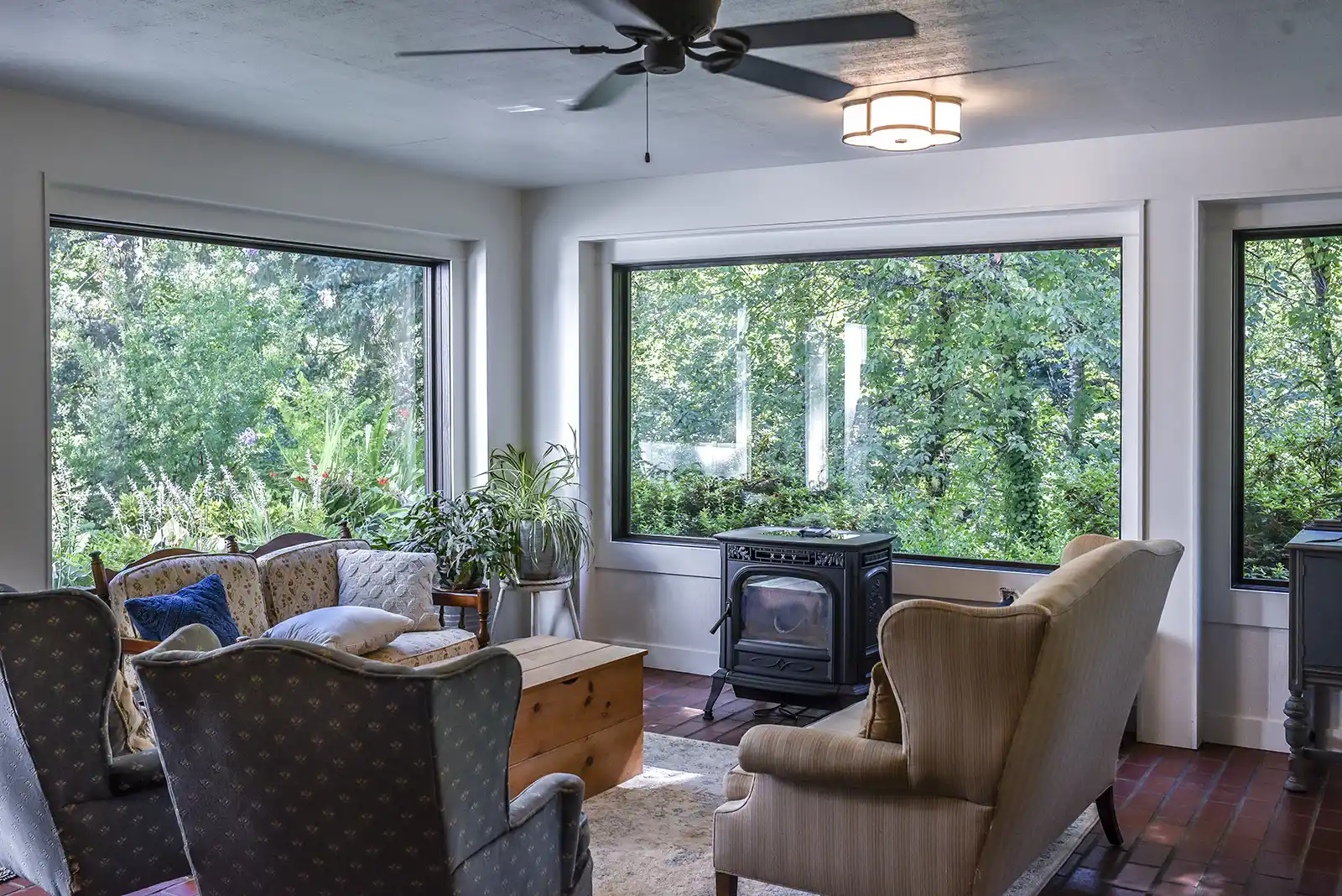 Living space with large windows showing lush greenery, vintage chairs, a wood stove, and potted plants.
