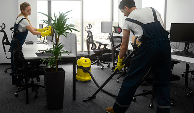 Two cleaners in uniforms cleaning an office with vacuum and wipes near desks and plants.