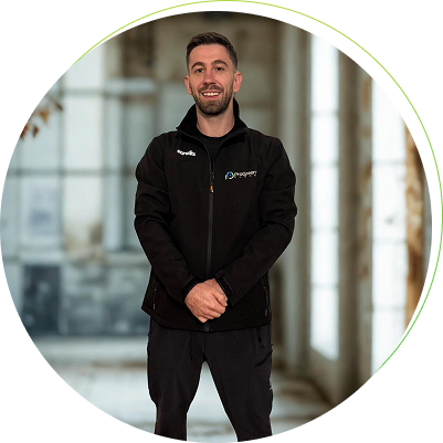 Man smiling in black jacket with logo, standing in a bright, spacious hallway with tall windows.