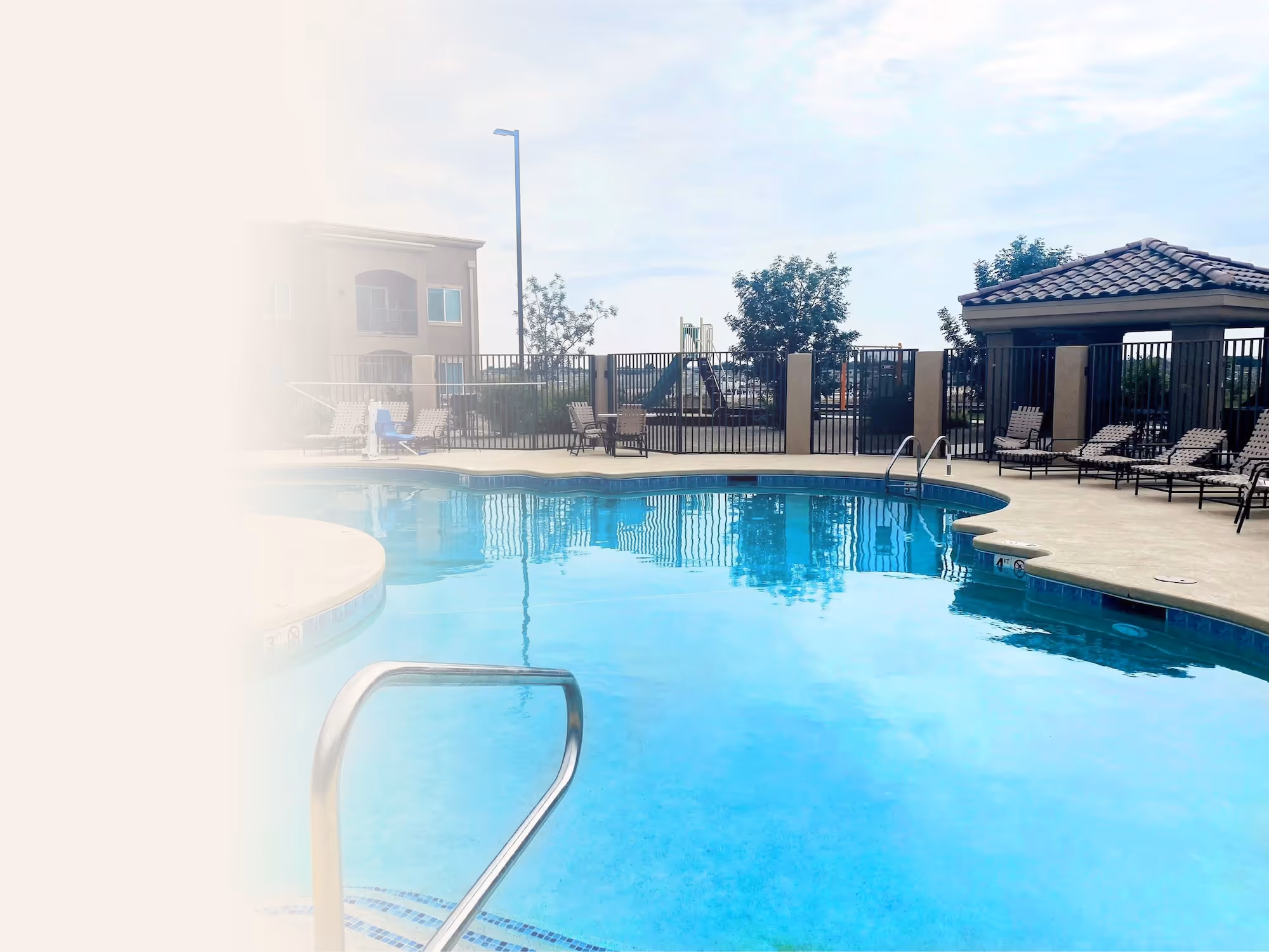 Outdoor swimming pool with lounge chairs and a building in the background under a cloudy sky.