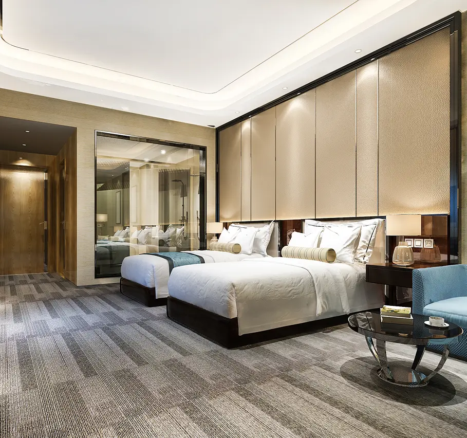 Modern hotel room with two neatly made beds featuring white linens and beige bolster pillows, a large padded headboard, bedside tables with lamps, a glass wall revealing a bathroom, a wooden door entrance, and a small round glass table next to a blue upholstered chair on a patterned carpet floor.