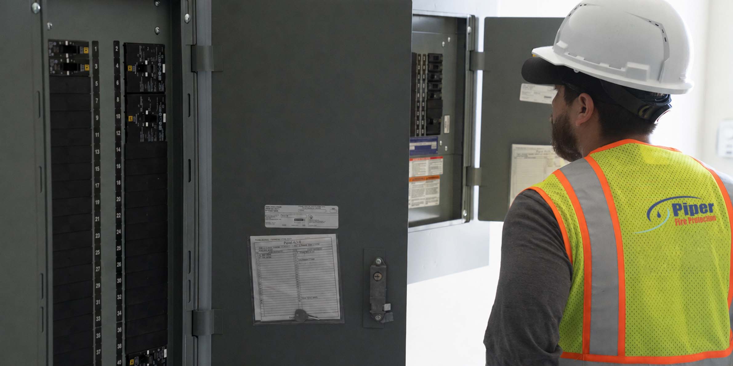 Man wearing a white hard hat and yellow safety vest with Piper Fire Protection logo inspecting an open electrical panel with circuit breakers.
