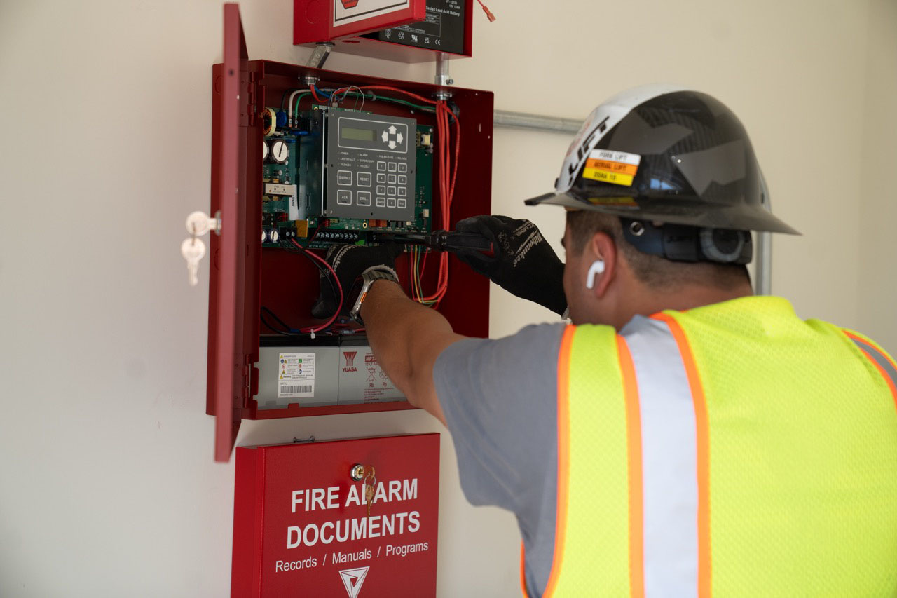 Technician wearing a safety vest, hard hat, gloves, and an earbud performing maintenance on an open fire alarm control panel mounted on a wall with a red locked box labeled 'FIRE ALARM DOCUMENTS' below it.