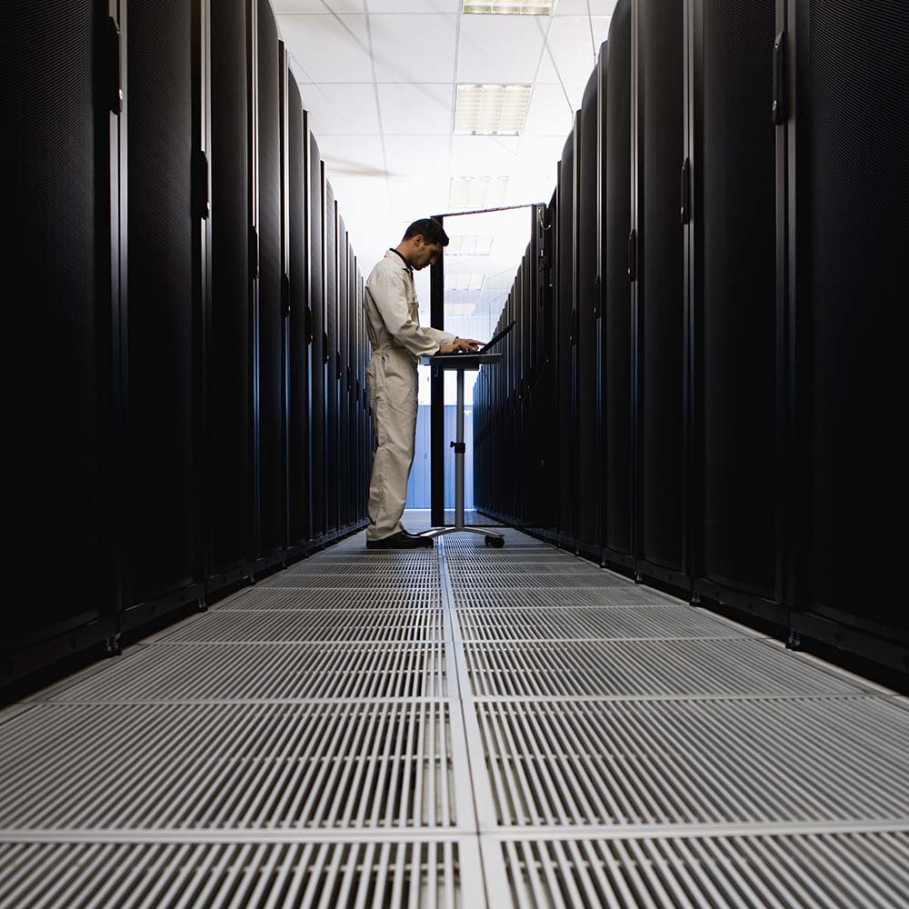 Man in light-colored coveralls working on a laptop on a stand in a corridor between rows of tall black server racks in a data center.