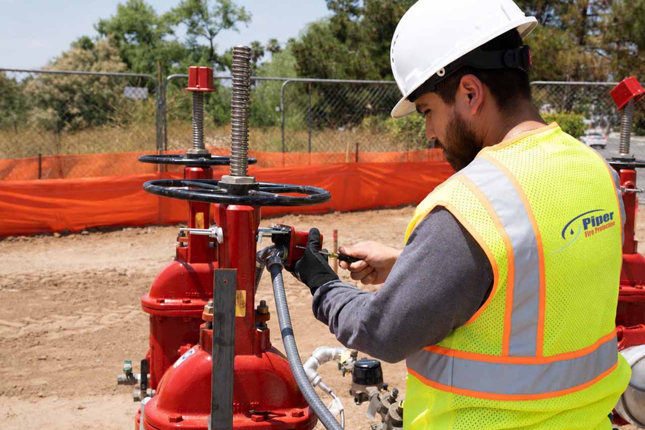 A construction worker wearing a white hard hat and yellow safety vest with the logo 'Piper Fire Protection' adjusts a valve on red industrial equipment outdoors at a worksite with orange safety fencing and trees in the background.