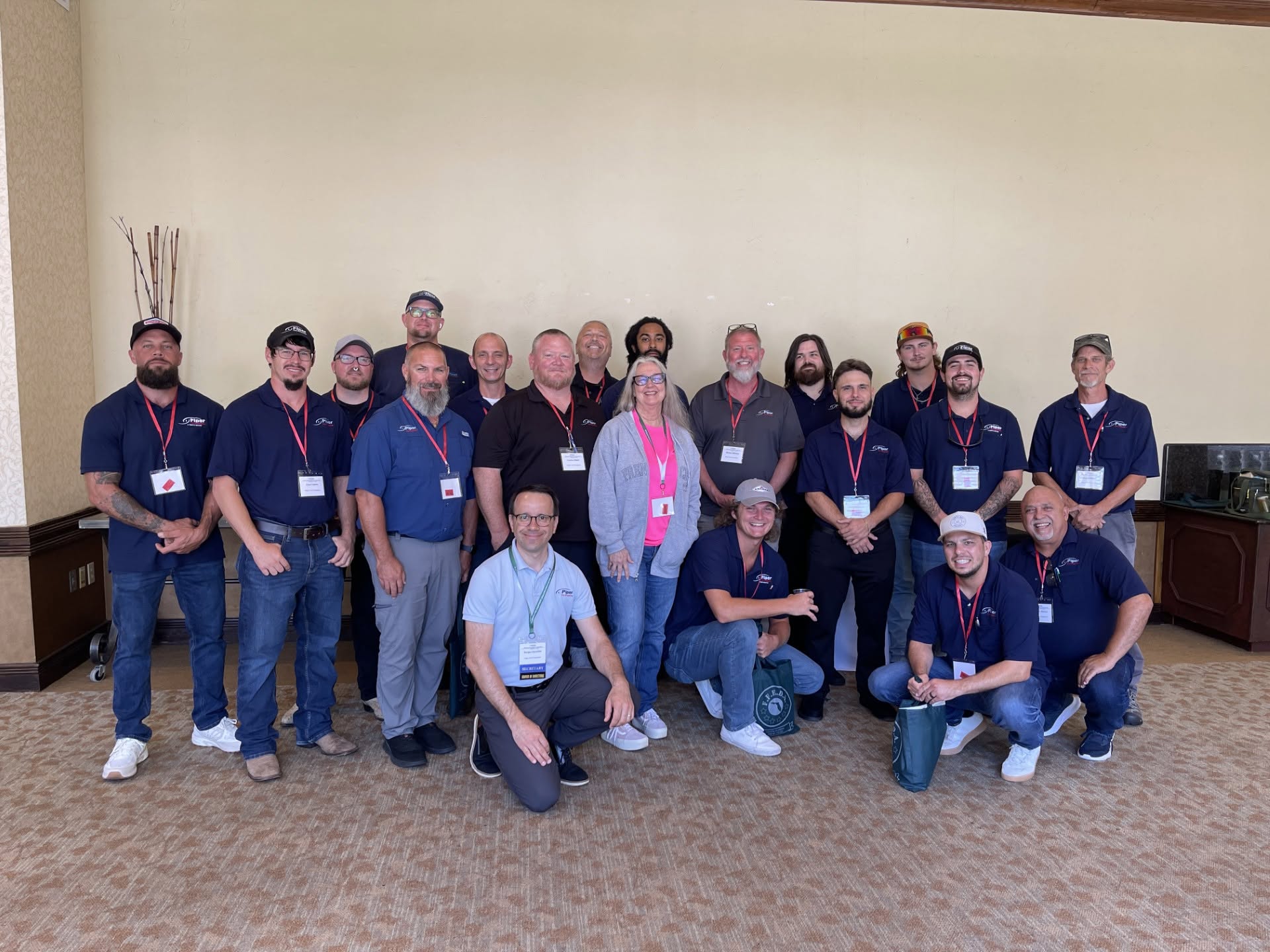Group of 19 people posing indoors against a beige wall, mostly men wearing navy blue shirts with logos and red lanyards, with two men kneeling in front holding bags and one woman in a gray hoodie near the center.