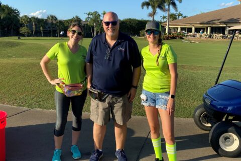 Two women and one man standing on a golf course near a golf cart, smiling at the camera on a sunny day; the women wear bright green shirts, and the man wears a dark blue polo.