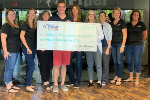 Eight people standing indoors on a wooden floor holding a large check made out to Children's Pain Foundation for $10,276.86, dated 9-23-19, from Piper Fire Protection.