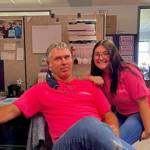 Two people in an office setting wearing matching bright pink shirts; one is sitting and the other is leaning on the chair, smiling.