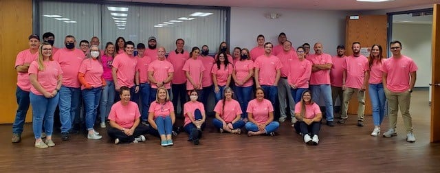 Group of 32 people wearing pink shirts standing and sitting in a large room with wooden floor and beige walls, some wearing face masks.