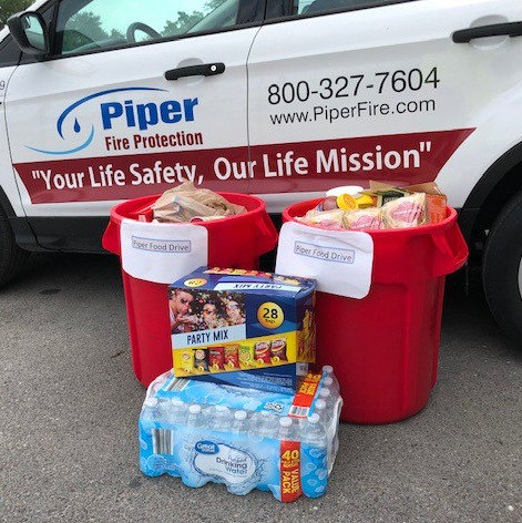Two red bins labeled 'Piper Food Drive' filled with various food items, next to a pack of bottled water and a party mix snack box, positioned in front of a white vehicle with Piper Fire Protection branding and the slogan 'Your Life Safety, Our Life Mission.'