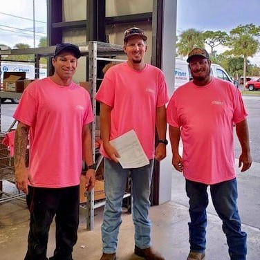 Three men wearing matching bright pink t-shirts standing inside a warehouse area near an open garage door with pallets and boxes in the background. The man in the center holds a sheet of paper. Outside the open door, trees and vehicles are visible under a partly cloudy sky.