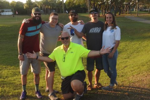 Group of six adults smiling outdoors on grass in a park with trees in the background, one man in a bright yellow shirt kneeling in front with arms open, others standing behind him.