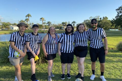 Six people standing outdoors on green grass under a clear blue sky, five of them wearing black and white striped referee shirts, and one in a sleeveless striped top, posing and smiling at the camera.
