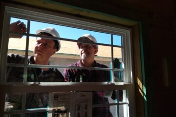 Two construction workers wearing white hard hats install or inspect a window from outside a building during daylight.