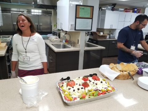 A woman wearing a white shirt and striped skirt laughs while standing behind a kitchen counter with a large salad topped with tomatoes and olives, a container of dressing, and a basket of bread rolls. A man wearing glasses and gloves works nearby in the background.