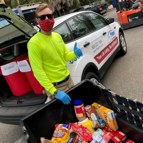 Man wearing sunglasses, a red face mask, blue gloves, and a bright yellow shirt giving a thumbs-up next to a vehicle with open trunk filled with red coolers, standing behind a container filled with various grocery items including peanut butter, cereal, and snacks.