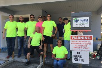 Group of nine people, most wearing bright yellow-green shirts, posing at a construction site next to a sign for Habitat for Humanity warning against trespassing and dumping, with the project address number 607 visible.