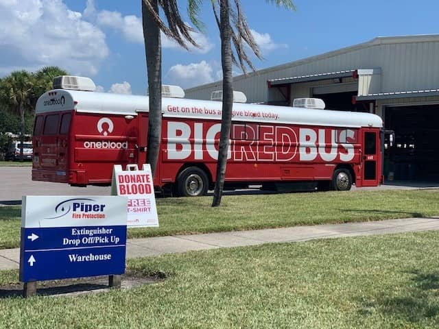 Two blood donation buses labeled ‘oneblood’ and ‘BIG RED BUS’ parked near a warehouse building with a sign for Piper Fire Protection indicating extinguisher drop off and warehouse directions. A smaller sign reads ‘DONATE BLOOD FREE T-SHIRT’.