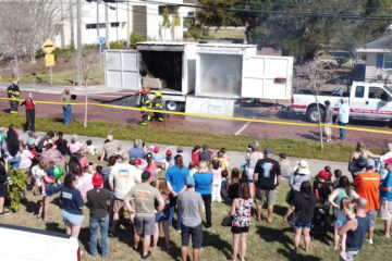 Crowd of people gathered on a lawn watching firefighters operate at the back of a firetruck, with yellow caution tape cordoning off the area.