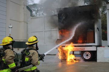 Two firefighters in full gear and helmets are spraying water to extinguish a fire on a large industrial container emitting thick black smoke and flames.