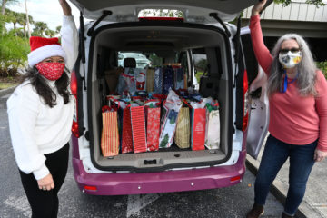 Two women wearing festive face masks stand by the open back of a vehicle filled with wrapped holiday gifts, one woman wears a Santa hat and white sweater and the other a red sweater and glasses.