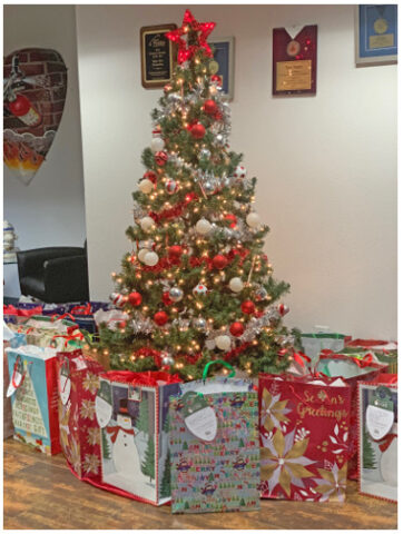 Decorated Christmas tree with red and gold ornaments and lights, topped with a red star, surrounded by colorful gift bags on a wooden floor in an indoor setting with framed awards on the wall.