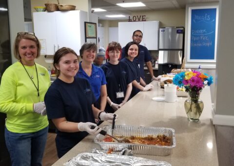 Six smiling volunteers standing behind a counter in a kitchen, serving food from aluminum trays, with a vase of colorful flowers on the counter and a sign reading 'LOVE' in the background.